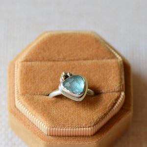 May include: A silver ring featuring a faceted, light blue, triangular-shaped gemstone set in a bezel. The setting includes small, decorative gold-toned metal spheres clustered at the top edge of the stone, attached to a smooth, polished silver band.