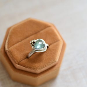 May include: A silver ring featuring a faceted, light blue-green stone in a triangular setting. The band is smooth and metallic, with three small, round gold-toned beads clustered on one side of the stone, creating a distinct, asymmetrical design.