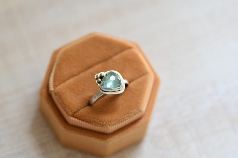 May include: A silver ring featuring a faceted, light blue-green stone in a triangular setting. The band is smooth and metallic, with three small, round gold-toned beads clustered on one side of the stone, creating a distinct, asymmetrical design.