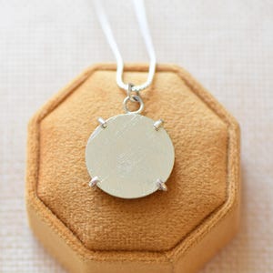May include: A silver-toned circular pendant necklace featuring a brushed metal finish. The round disc is secured by four small prongs and hangs from a thin, delicate silver-coloured chain. The pendant has a flat, matte surface texture.