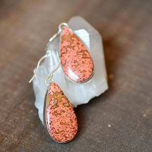 May include: A pair of teardrop-shaped earrings featuring a speckled peach and brown pattern set in silver-toned metal. Each earring hangs from a simple silver-toned hook, showcasing a smooth, polished surface with a distinct organic texture.