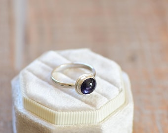May include: A silver ring with a bezel setting featuring a dark purple gemstone. The ring is displayed on a white velvet ring box.