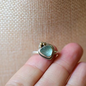 May include: A silver ring featuring a single, irregular, light blue, translucent stone set in a bezel. The setting includes three small, round, gold-coloured metal accents clustered at the top edge of the stone, mounted on a simple silver band.