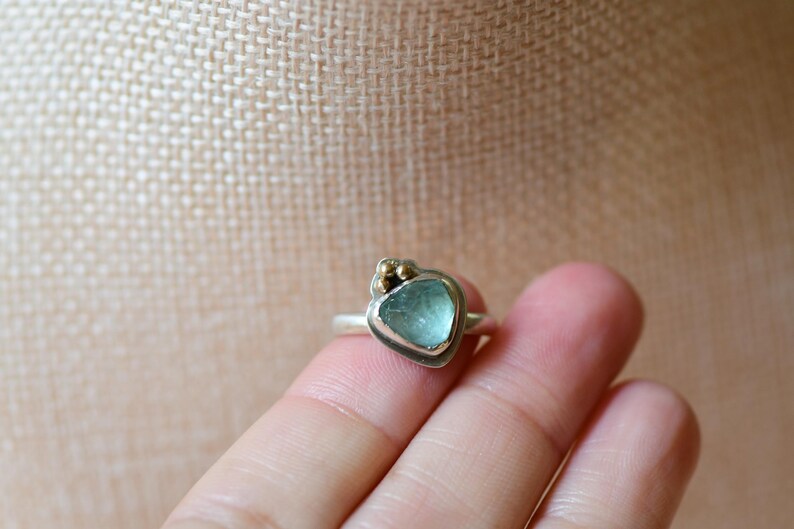 May include: A silver ring featuring a single, irregular, light blue, translucent stone set in a bezel. The setting includes three small, round, gold-coloured metal accents clustered at the top edge of the stone, mounted on a simple silver band.