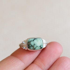 May include: A silver ring with a rectangular, light green stone with dark green veining. The ring has a feather design on each side of the stone. The ring is shown on a person's finger.