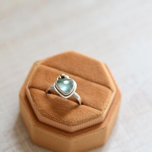 May include: A silver ring featuring a faceted, triangular light blue gemstone set in a bezel. The ring band is smooth and metallic, with a small gold-toned accent bead positioned directly above the top point of the triangular stone setting.