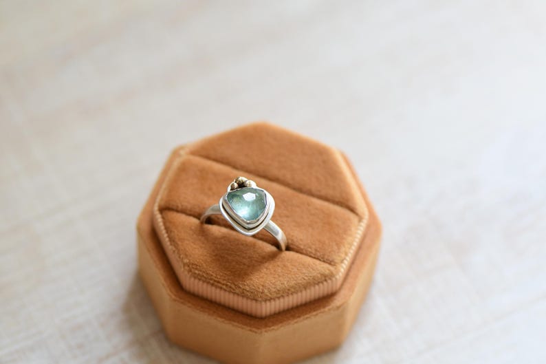 May include: A silver ring featuring a faceted, triangular light blue gemstone set in a bezel. The ring band is smooth and metallic, with a small gold-toned accent bead positioned directly above the top point of the triangular stone setting.
