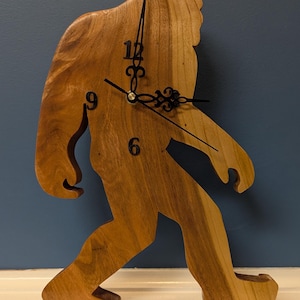 May include: A wall clock shaped like a walking Bigfoot figure, crafted from light brown wood with a visible natural grain. The clock features black metal hands and black numerals for 12, 9, and 6 positioned on the torso of the wooden figure.