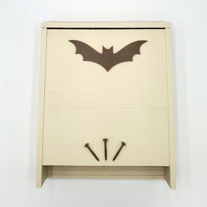 May include: A cream-colored wooden bat house with a brown bat silhouette on the front. Three decorative nails are placed below the opening. The bat house is designed to provide shelter for bats.