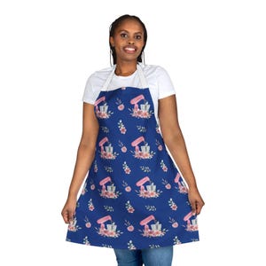 May include: A blue apron with a pink and white floral pattern featuring a kitchen mixer. The apron has adjustable straps and a large pocket.