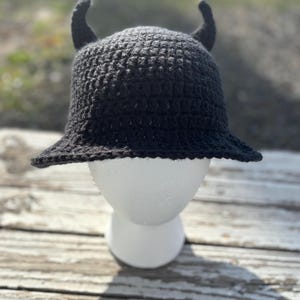 May include: A black crochet hat with two curved horns on top. The hat has a wide brim and is displayed on a white mannequin head. The hat is made of a dark, textured yarn and appears handmade. The background is blurred, with natural light.