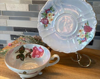 May include: A vintage teacup and saucer set with a floral design. The teacup has a single pink rose and green leaves on a light blue background. The saucer has a more elaborate floral design with pink, yellow, and white flowers on a light blue background.