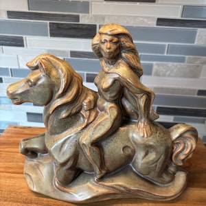 Puede incluir: Una escultura de cerámica que muestra una figura con cabello largo y fluido montada sobre un caballo. La pieza tiene un acabado de esmalte moteado en tonos tierra marrón y verde con una textura suave, representando a la figura sobre el caballo.
