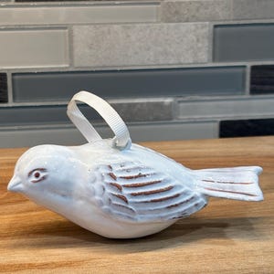 May include: White ceramic bird ornament with a brown glaze detail. The bird has a loop for hanging and a small red eye.