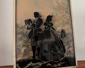 May include: A framed silhouette art piece depicting a man and woman in period clothing. The black silhouettes are set against a soft, colorful background with a gold-colored frame. The artwork is displayed on a gold-colored stand.