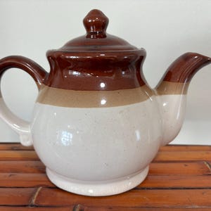 Puede incluir: Una tetera de cerámica con tapa, asa y boquilla. La tetera tiene un cuerpo redondeado con una base blanca, una sección media beige y una parte superior marrón oscuro. El asa y la boquilla también son marrones y beige.
