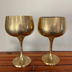 May include: Two vintage gold-colored goblets with etched floral designs. The goblets have a rounded bowl shape, slender stems, and wide bases. The goblets are made of a shiny, reflective metal, and are displayed on a wooden surface.