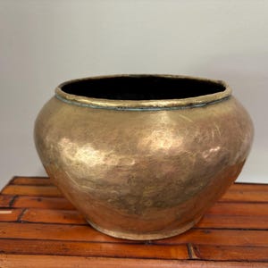 May include: A hammered brass bowl with a wide, rounded body and a dark interior. The bowl sits on a bamboo surface. The brass has a warm, golden hue, and the rim is slightly lighter in color.
