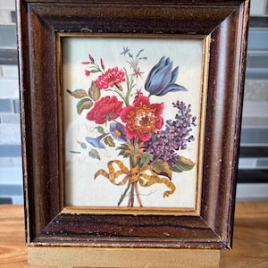 Pode incluir: Uma impressão floral emoldurada com um buquê de flores vermelhas, roxas e azuis, amarradas com uma fita amarela. A moldura é marrom escura com uma borda dourada e é exibida em um pequeno cavalete.