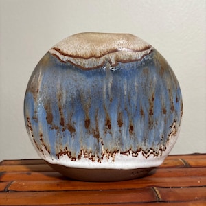 May include: A round ceramic vase with a wide opening. The top is tan, transitioning to a blue center with brown streaks, and a white base. The vase sits on a bamboo surface.