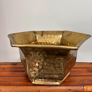 May include: A brass planter with a hammered texture and a geometric design. The planter has a wide opening and a surface with small, rounded indentations. The brass has a warm, golden colour, and the planter sits on a bamboo surface.
