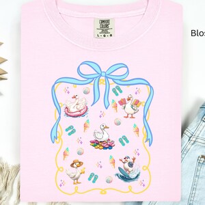 May include: A pink t-shirt with a blue ribbon and a graphic of ducks in a summer scene. The graphic includes ducks on a raft, ducks on a surfboard, and ducks with ice cream cones. The text "COMFORT COLORS" is on the tag.