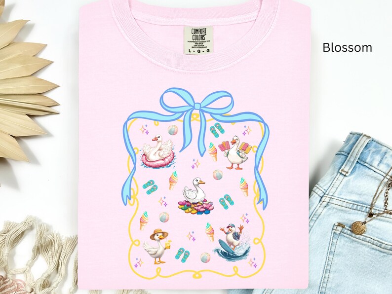 May include: A pink t-shirt with a blue ribbon and a graphic of ducks in a summer scene. The graphic includes ducks on a raft, ducks on a surfboard, and ducks with ice cream cones. The text "COMFORT COLORS" is on the tag.