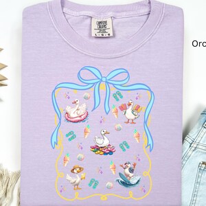 May include: A lavender t-shirt with a graphic of four ducks enjoying summer activities. The graphic is surrounded by a blue ribbon with a bow and a gold frame. The text "COMFORT COLORS" is printed on the tag.