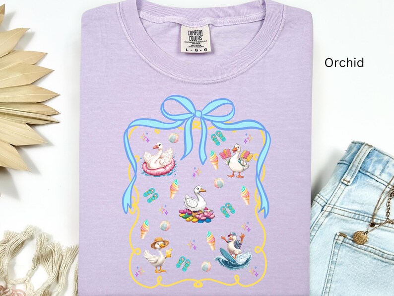 May include: A lavender t-shirt with a graphic of four ducks enjoying summer activities. The graphic is surrounded by a blue ribbon with a bow and a gold frame. The text "COMFORT COLORS" is printed on the tag.