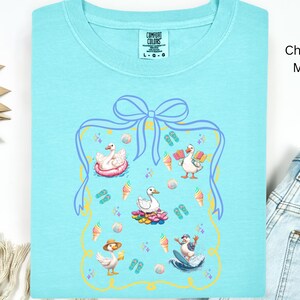 May include: A light blue t-shirt with a graphic of ducks enjoying summer activities. The graphic is surrounded by a blue and yellow frame with a bow at the top. The text "COMFORT COLORS" and "L-G-G" is visible on the shirt. The shirt is labeled "Chalky Mint".