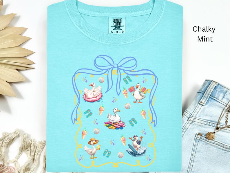 May include: A light blue t-shirt with a graphic of ducks enjoying summer activities. The graphic is surrounded by a blue and yellow frame with a bow at the top. The text "COMFORT COLORS" and "L-G-G" is visible on the shirt. The shirt is labeled "Chalky Mint".