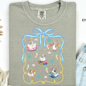 May include: Khaki t-shirt with a blue bow and a framed design featuring five ducks in various summer activities. The ducks are wearing hats and sunglasses and are surrounded by ice cream cones, beach balls, and other summer-themed items.