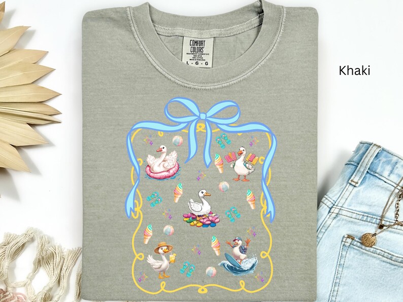 May include: Khaki t-shirt with a blue bow and a framed design featuring five ducks in various summer activities. The ducks are wearing hats and sunglasses and are surrounded by ice cream cones, beach balls, and other summer-themed items.