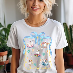 May include: White t-shirt with a blue ribbon and a graphic of ducks enjoying summer activities, including ice cream cones, flip flops, and a surfboard.