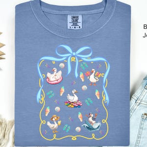 May include: A blue jean t-shirt with a graphic of a blue ribbon and four white ducks with summer accessories. The text "COMFORT COLORS" is printed on the shirt.