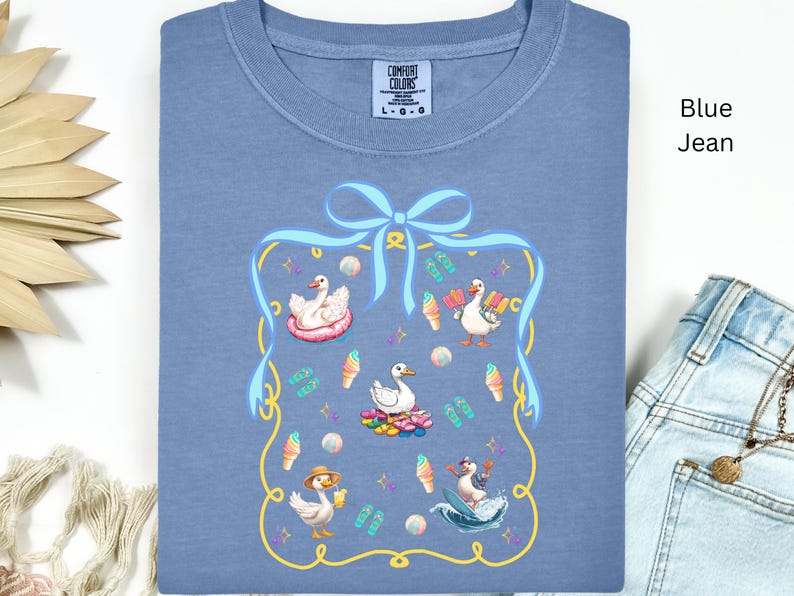 May include: A blue jean t-shirt with a graphic of a blue ribbon and four white ducks with summer accessories. The text "COMFORT COLORS" is printed on the shirt.