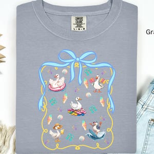 May include: A gray t-shirt with a blue ribbon and a yellow frame. Inside the frame are four cartoon ducks, two with ice cream cones, one in a pool float, and one surfing. The text "Comfort Colors" is on the shirt.