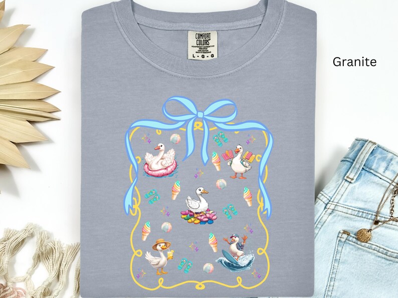 May include: A gray t-shirt with a blue ribbon and a yellow frame. Inside the frame are four cartoon ducks, two with ice cream cones, one in a pool float, and one surfing. The text "Comfort Colors" is on the shirt.