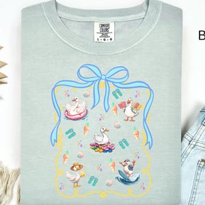 May include: A light blue t-shirt with a graphic of a blue ribbon and a frame with various images of ducks enjoying summer activities, including swimming, sunbathing, and surfing. The text "COMFORT COLORS" and "L-G-O" are visible on the shirt.