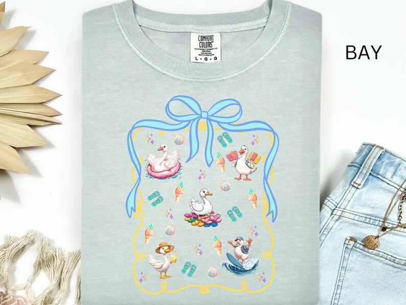 May include: A light blue t-shirt with a graphic of a blue ribbon and a frame with various images of ducks enjoying summer activities, including swimming, sunbathing, and surfing. The text "COMFORT COLORS" and "L-G-O" are visible on the shirt.