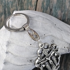 May include: Silver-toned metal keychain featuring a religious emblem. The emblem depicts a crucifix with figures and ornate detailing. The keychain has a split ring and a decorative connector.