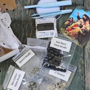 May include: A DIY rosary kit containing black 8mm Hail Mary beads, grey 8mm Our Father beads, silver spacer beads, a metal crucifix, a metal center piece, a spool of Nylon Power 0.90mm string, light blue tweezers, and a religious prayer card.