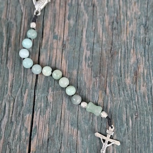 May include: A single-decade rosary featuring light green, smooth, round beads strung on a black cord. The rosary includes silver-toned metal spacers, a silver-toned crucifix with the letters INRI, and a silver-toned lobster claw clasp.