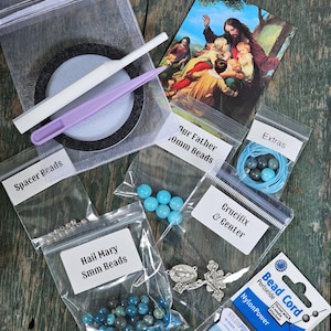 May include: A DIY rosary making kit containing a white and a purple plastic tool, a religious card, and several clear plastic bags labeled Spacer Beads, Our Father 10mm Beads, Extras, Hail Mary 8mm Beads, and Crucifix & Center, plus a spool of blue nylon bead cord.