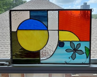 May include: Stained glass panel with a geometric design. The panel features a blue circle over a yellow half-circle, a red square, and a blue and white flower on a light blue background. The panel is framed in black.