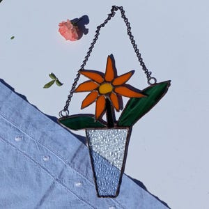Op de afbeelding: Een zonnenbloem van gekleurd glas in een witte vaas met een zilveren glitterafwerking. De bloem is oranje met een gele kern en groene bladeren. De vaas hangt aan een zilveren ketting.