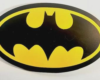 May include: An oval-shaped wall plaque featuring the iconic black bat silhouette centered against a vibrant yellow background, enclosed within a thick black border. The design is flat and graphic, displaying the classic superhero emblem.