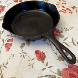 May include: A black cast iron frying pan with a long handle and a hanging loop. The pan is round with a slightly sloped side. The pan is on a floral tablecloth with red roses.