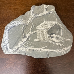 Puede incluir: Un grabado en pizarra gris de un pájaro en un comedero. El comedero es una pequeña estructura de madera con un tejado inclinado. El pájaro está posado en una rama, y el grabado incluye ramas de árboles y la firma del artista, "M.RICE."