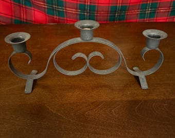 May include: A grey metal candle holder with three candle cups. The holder features a decorative, curved design with scrollwork in the centre. The candle holder is on a wooden surface with a red and green plaid background.